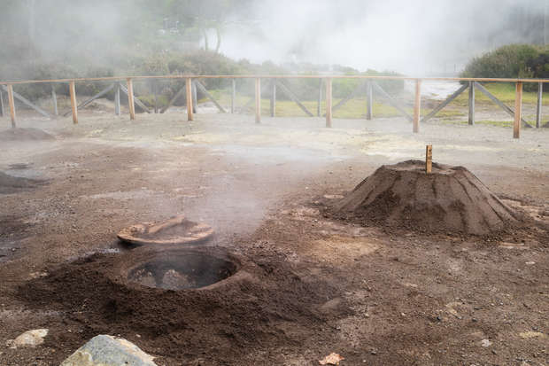 Steam rises from circular earthen mounds used for geothermal cooking. Wooden fences surround the area, with some greenery visible in the mist-covered background.